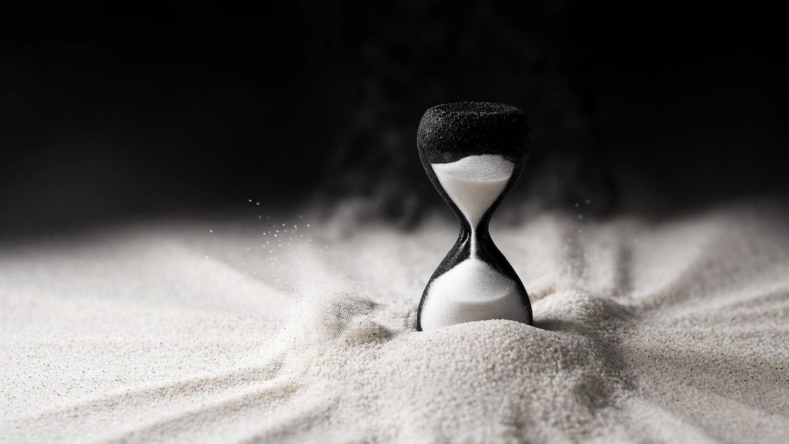 A black hourglass whose form is the sand, standing in a wide field of white sand under studio darkness.